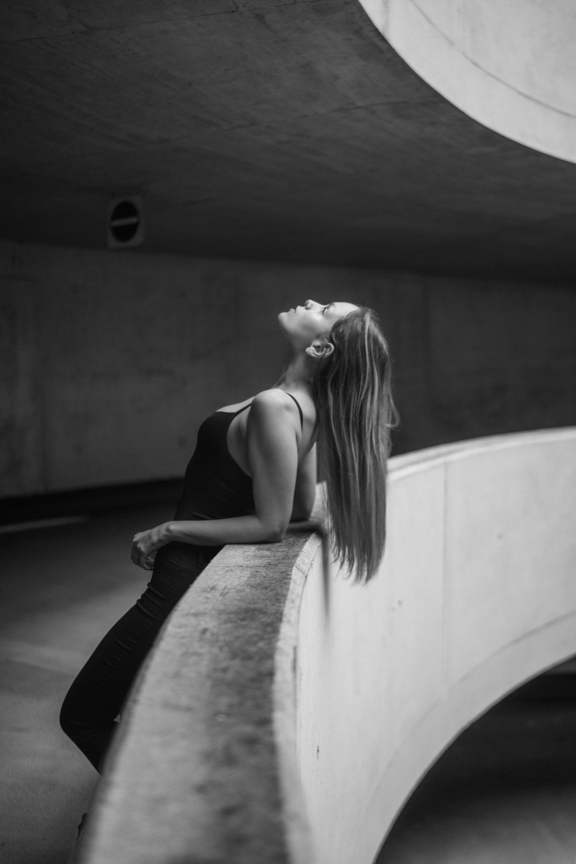 Frau lehnt sich entspannt an eine geschwungene Betonbrüstung in einem urbanen Parkhaus – Schwarz-Weiß-Portrait