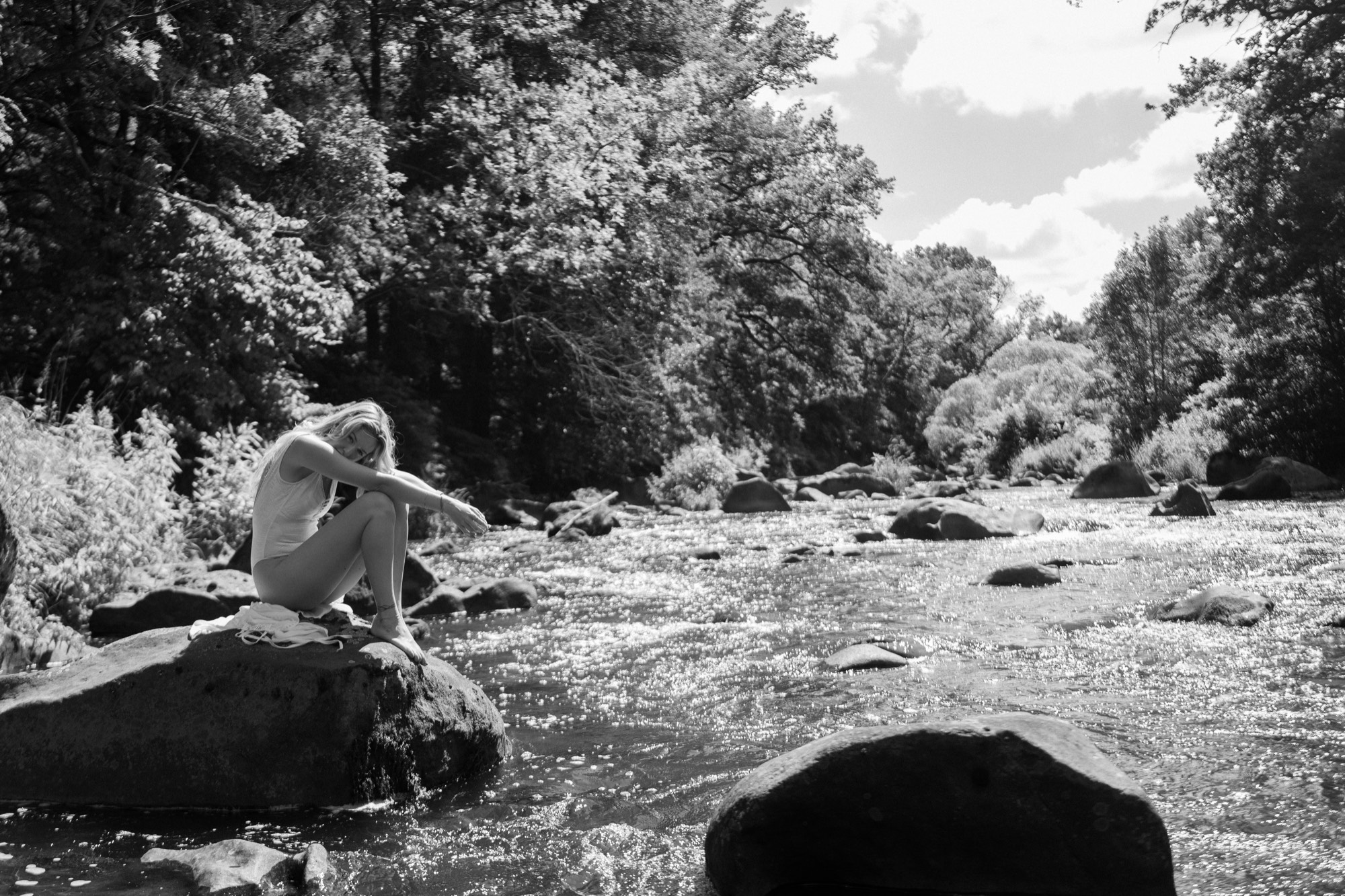 Frau sitzt entspannt auf einem Felsen in einem Fluss, umgeben von Wald – Schwarz-Weiß-Portrait aus einem Outdoor-Shooting